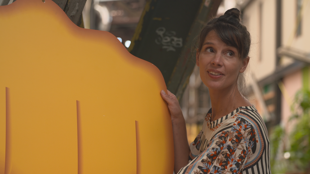 junge Frau mit hochgesteckter Friseur trägt eine riesige Pappe, die einen Teil eines Faust-Emojis zeigt