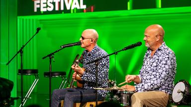 Die feisten sitzen auf Barhockern auf der Bühne. Vor ihnen stehen Mikrofonständer. Der Hintergrund ist grün beleuch