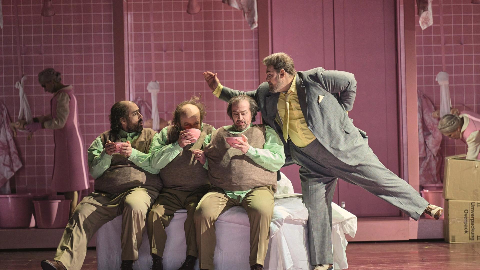 "Richard Strauss: Die Frau ohne Schatten": Drei Brüder (der Bucklige; der Einarmige, der Einäugige) sitzen nebeneinander. Alle halten eine rosafarbene Trinkschale in den Händen. Neben ihnen steht ein weiterer Mann auf einem Bein, mit einem Arm in Richtung der drei Brüder zeigend. Im Hintergrund ein ganz mit rosafarbenen Kacheln gefliester Raum und zwei Frauen mit rosa Kittelschürzen.