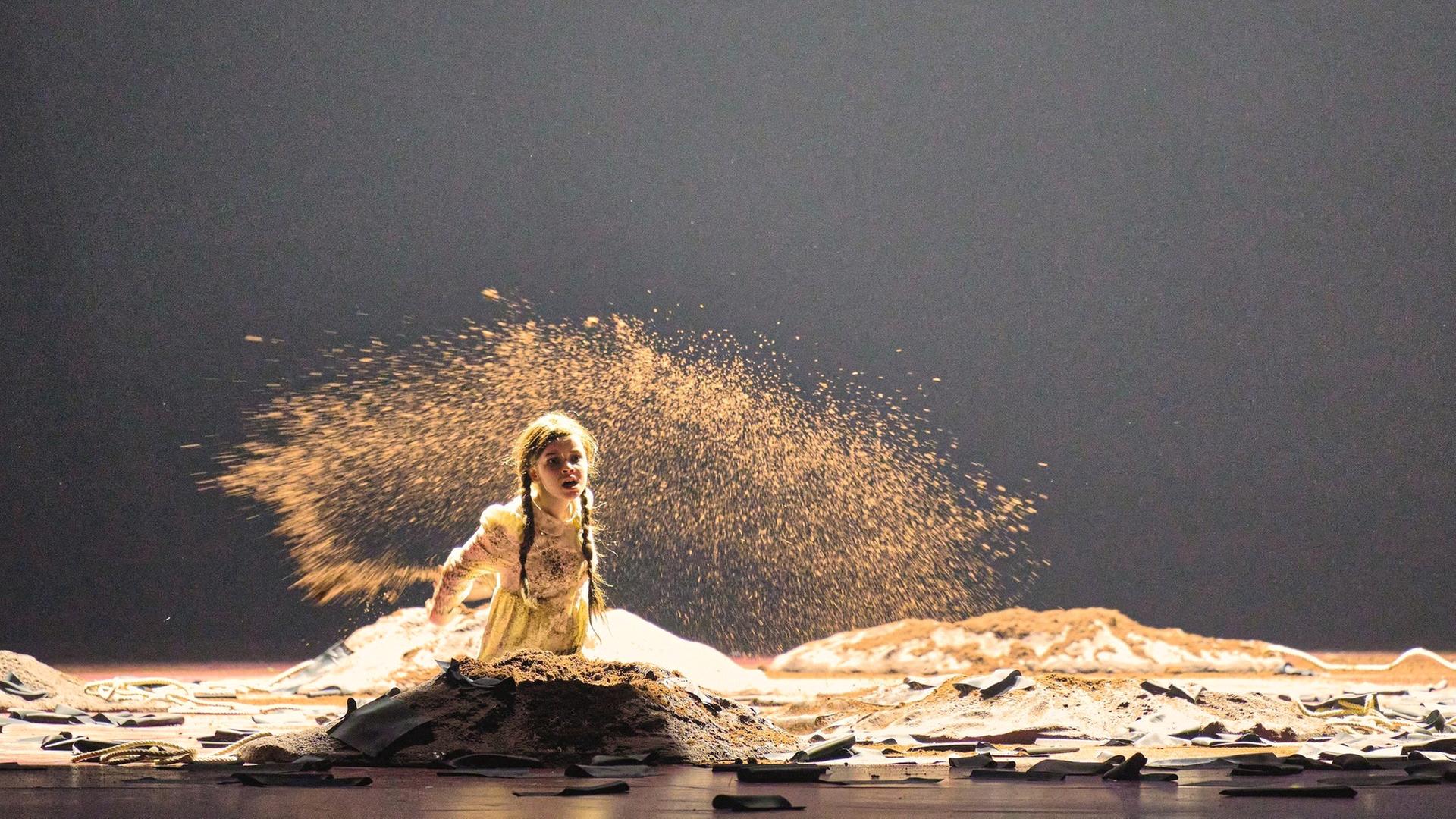 "Richard Strauss: Die Frau ohne Schatten": Ein Mädchen kniet auf dem Boden, vor ihr ein Haufen aus Sand und um sie herum weitere angedeutete Hügel. Sie trägt ein goldfarbenes Kleid und lange Zöpfe. Um sie herum scheint Sand zu fliegen. Alles ist in goldenen Farben gehalten.