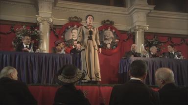 "Die glorreichen 10 - Die glorreichsten Gründer der Geschichte": Spielszene: Rosa Luxemburg steht auf einer Bühne. Vor ihr sitzt das Publikum, hinter ihr stehen zwei Tische, an denen Politiker und Politikerinnen sitzen. An der Wand hängen zwei Bilder von Ferdinand Lassalle und Karl Marx.