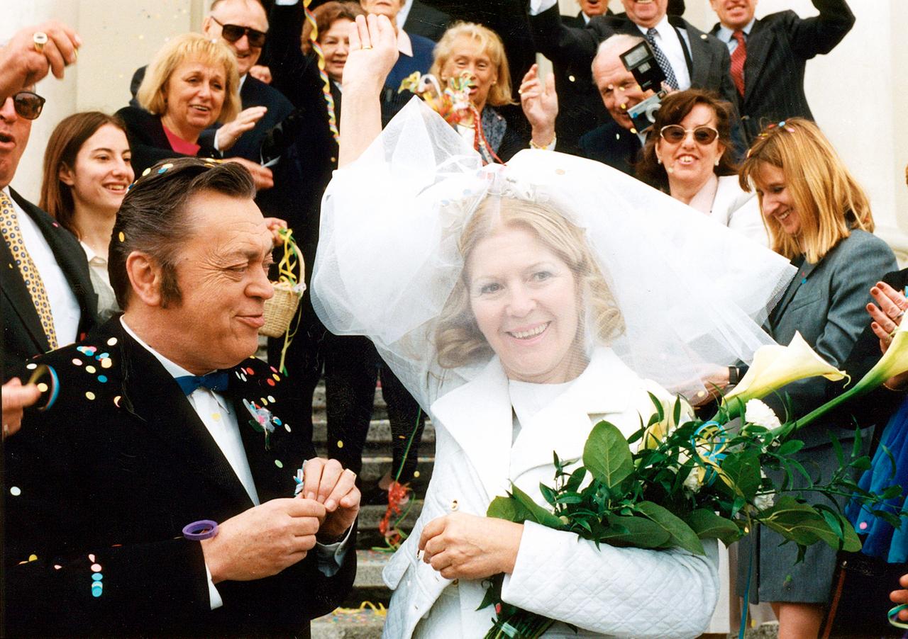 Das Bild zeigt eine festliche Szene vor einer Treppe, bei der eine Frau in einem Brautkleid mit einem Blumenstrauß strahlt. Neben ihr steht ein Mann im Anzug, während eine Menschenmenge feiert und Konfetti wirft.