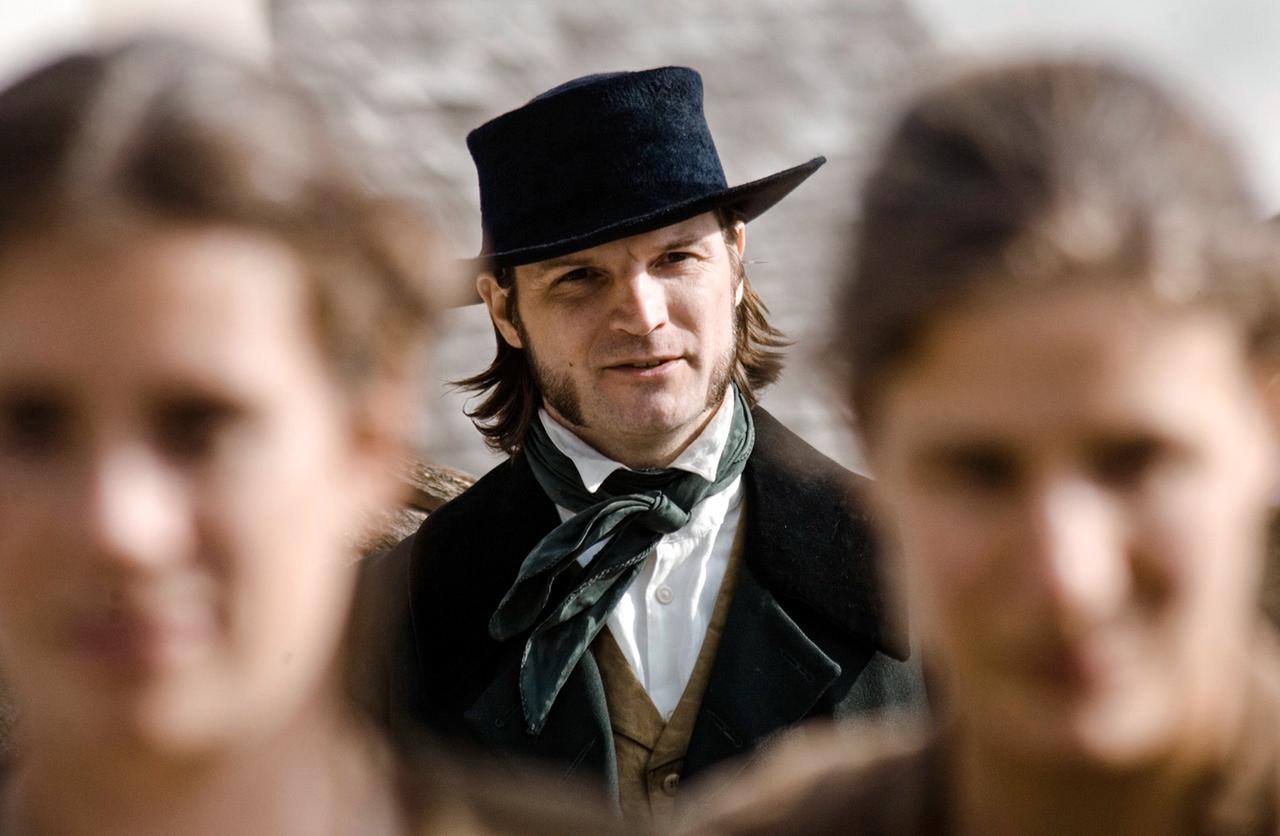 Ein Mann mit Hut sieht von hinten auf zwei Frauen vor ihm, die im Vordergrund zu sehen sind.