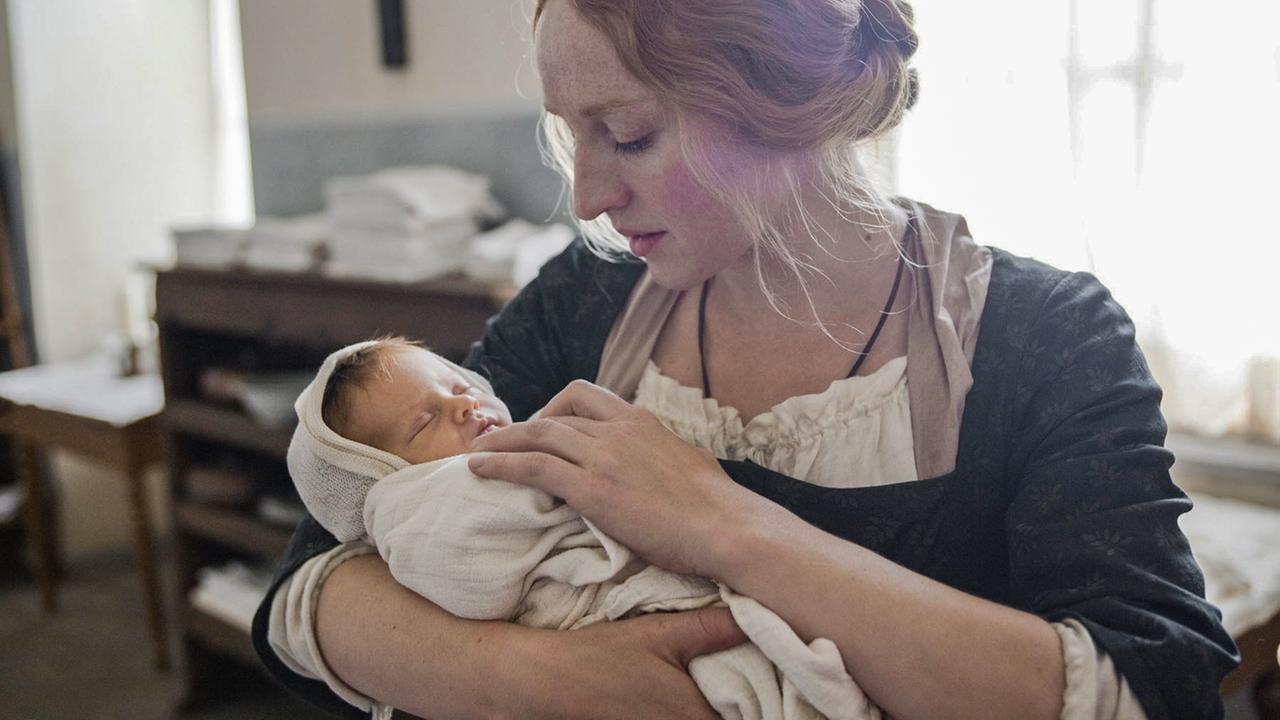 Ein junge Frau in historischer Kleidung hält ein Baby im Arm.