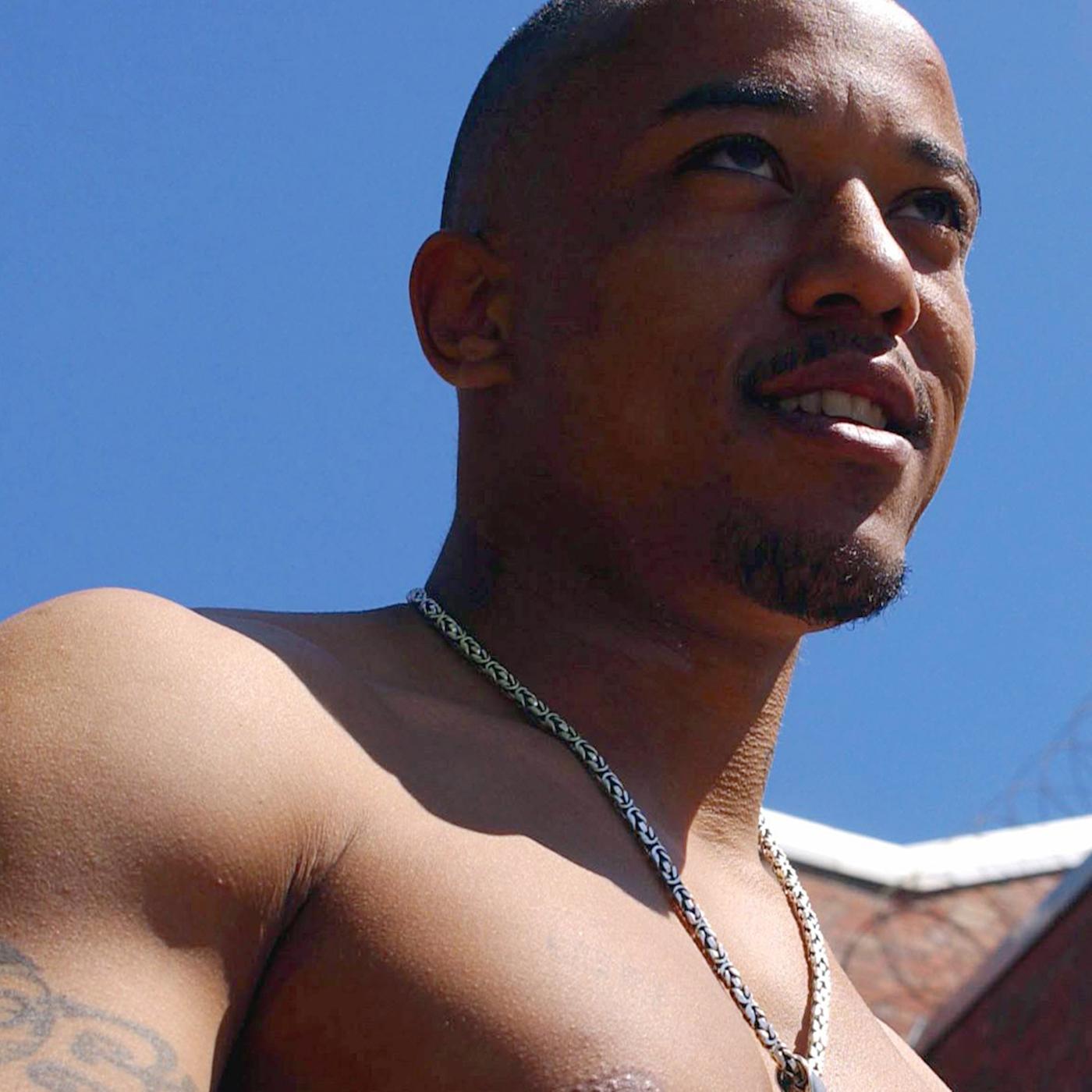 Nahaufnahme eines jungen Mannes mit nacktem Oberkörper. Im Hintergrund Gefängnismauern mit Stacheldraht.