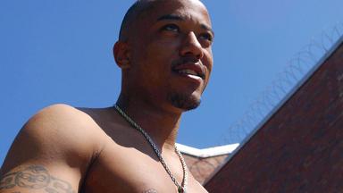 Nahaufnahme eines jungen Mannes mit nacktem Oberkörper. Im Hintergrund Gefängnismauern mit Stacheldraht.