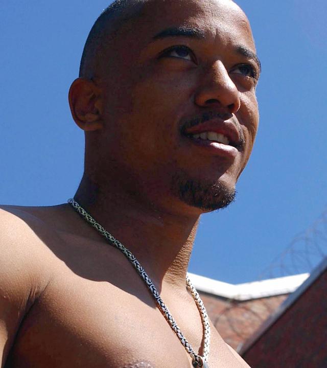 Nahaufnahme eines jungen Mannes mit nacktem Oberkörper. Im Hintergrund Gefängnismauern mit Stacheldraht.