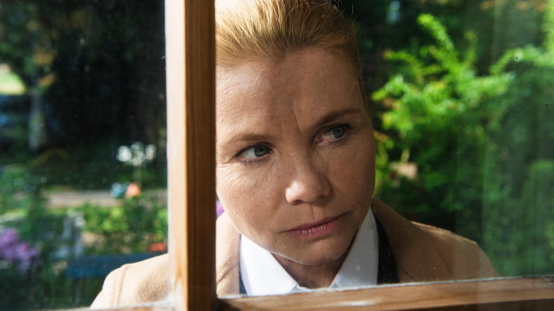 "Ella Schön - Die Inselbegabung": Ella (Annette Frier) schaut durch eine Fensterscheibe ins Innere eines Hauses. Hinter ihr sind verschwommene Ausschnitte eines Vorgartens erkennbar.