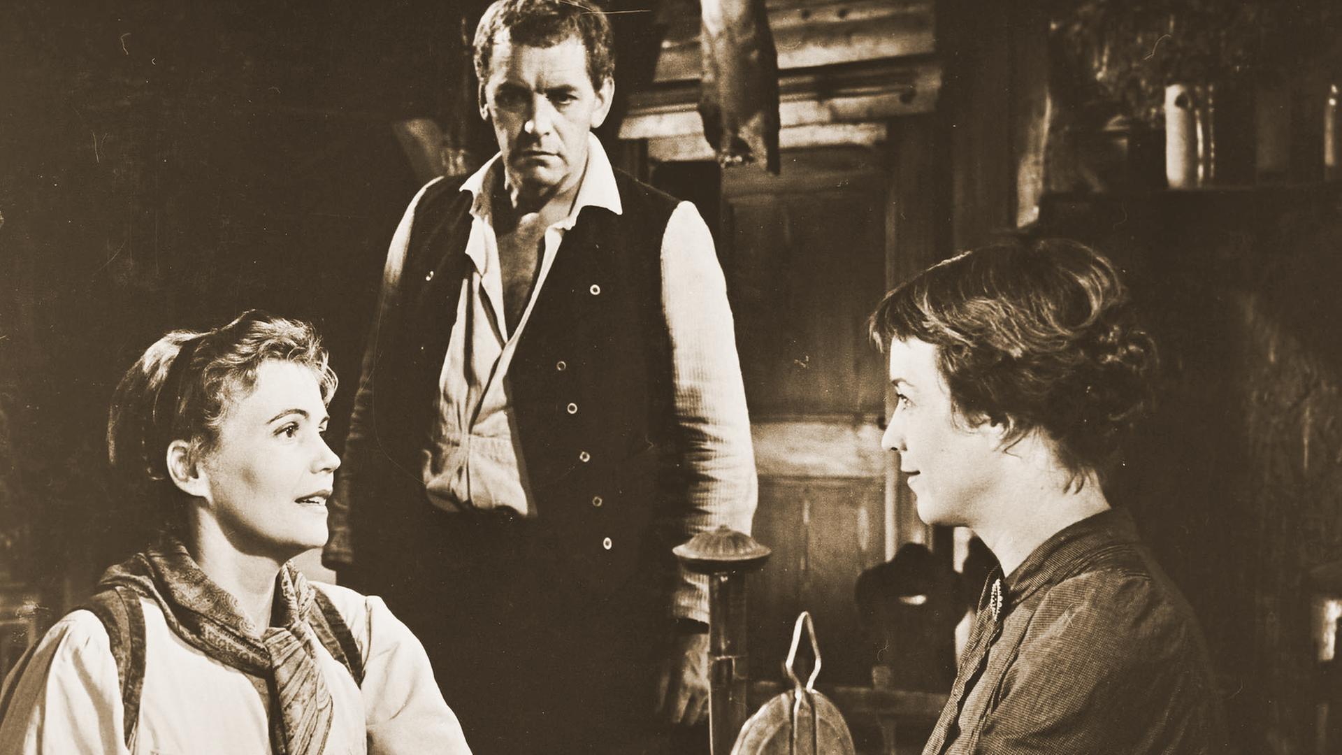 Zwei Frauen sitzen sich an einem Holztisch gegenüber und führen ein intensives Gespräch. Sie halten sich an den Händen. Im Hintergrund steht ein Mann mit mißtrauischem Blick. 