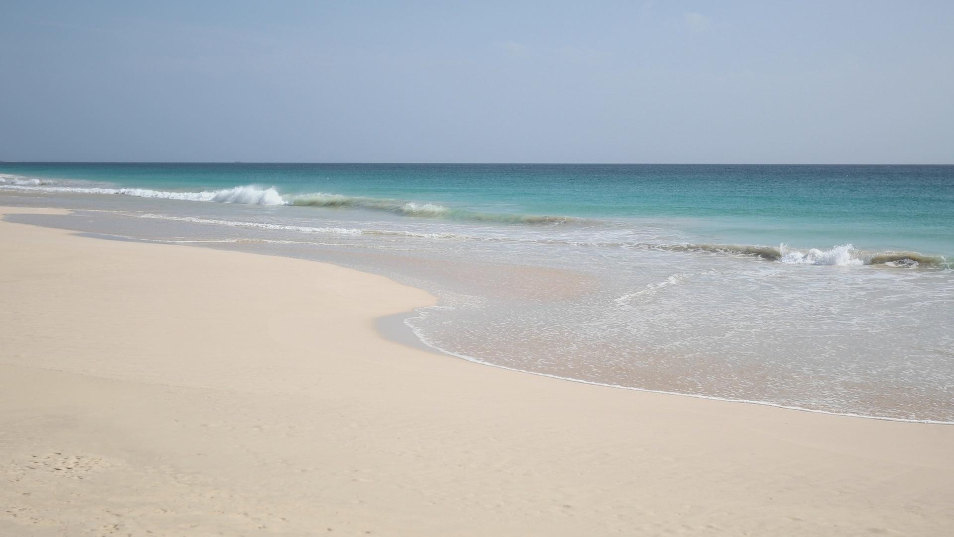Weißer Sandstrand unter blauem Himmel mit türkis-blauem Meer im Hintergrund.