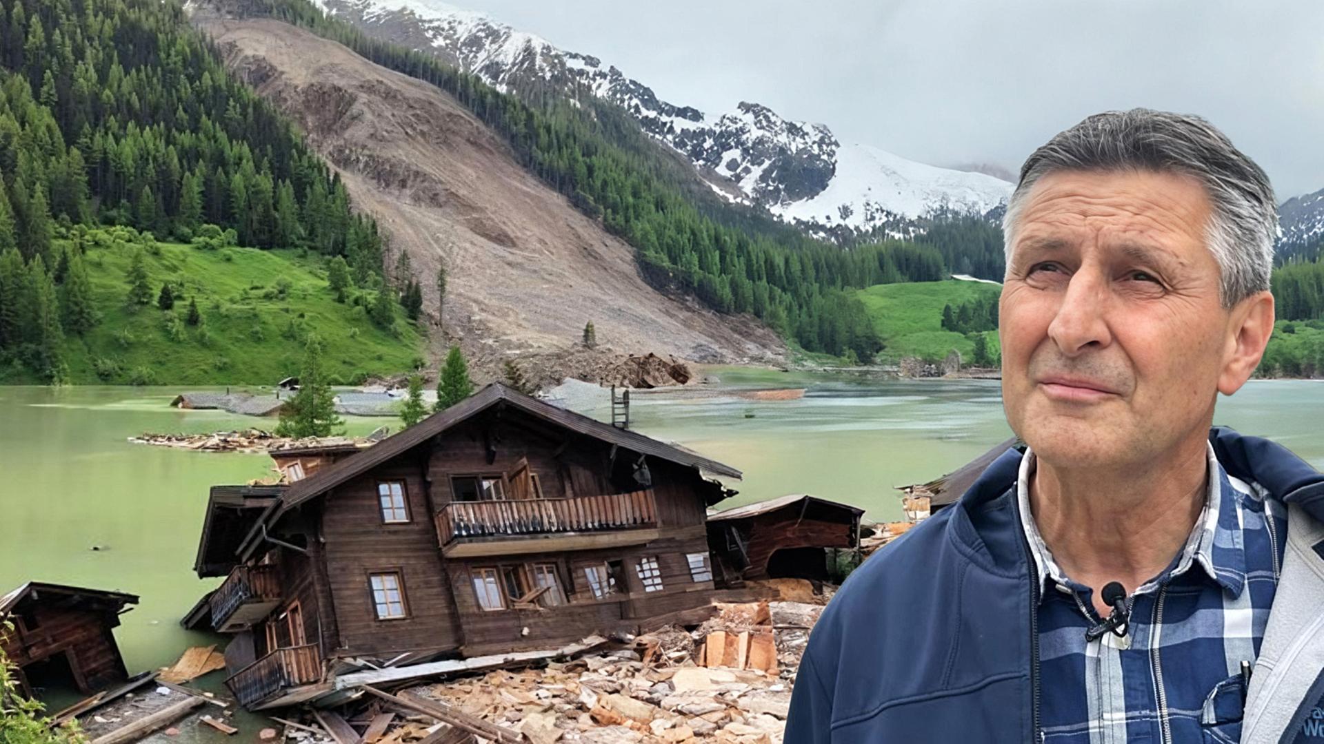 Ein Mann im karierten Hemd steht vor einem See mit überschwemmten Holzhäusern und einer großen Hangrutschung, dahinter ein bewaldeter Hang mit Schneebergen.