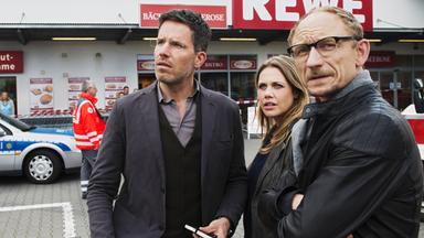 "Die Lebenden und die Toten (1) - Ein Taunuskrimi": Die Kommissare Oliver von Bodenstein (Tim Bergmann), Pia Kirchhoff (Felicitas Woll) und Kai Ostermann (Michael Schenk) stehen vor einem Supermarkt beisammen und schauen in die gleiche Richtung.