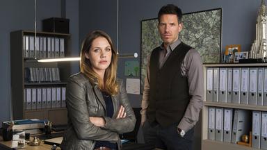 "Die Lebenden und die Toten (2) - Ein Taunuskrimi": Pia Kirchhoff (Felicitas Woll) lehnt mit vor der Brust verschränkten Armen an ihrem Schreibtisch im Büro. Hinter dem Schreibtisch steht Oliver von Bodenstein (Tim Bergmann), die Hände in den Hosentaschen. Beide schauen in die Kamera.