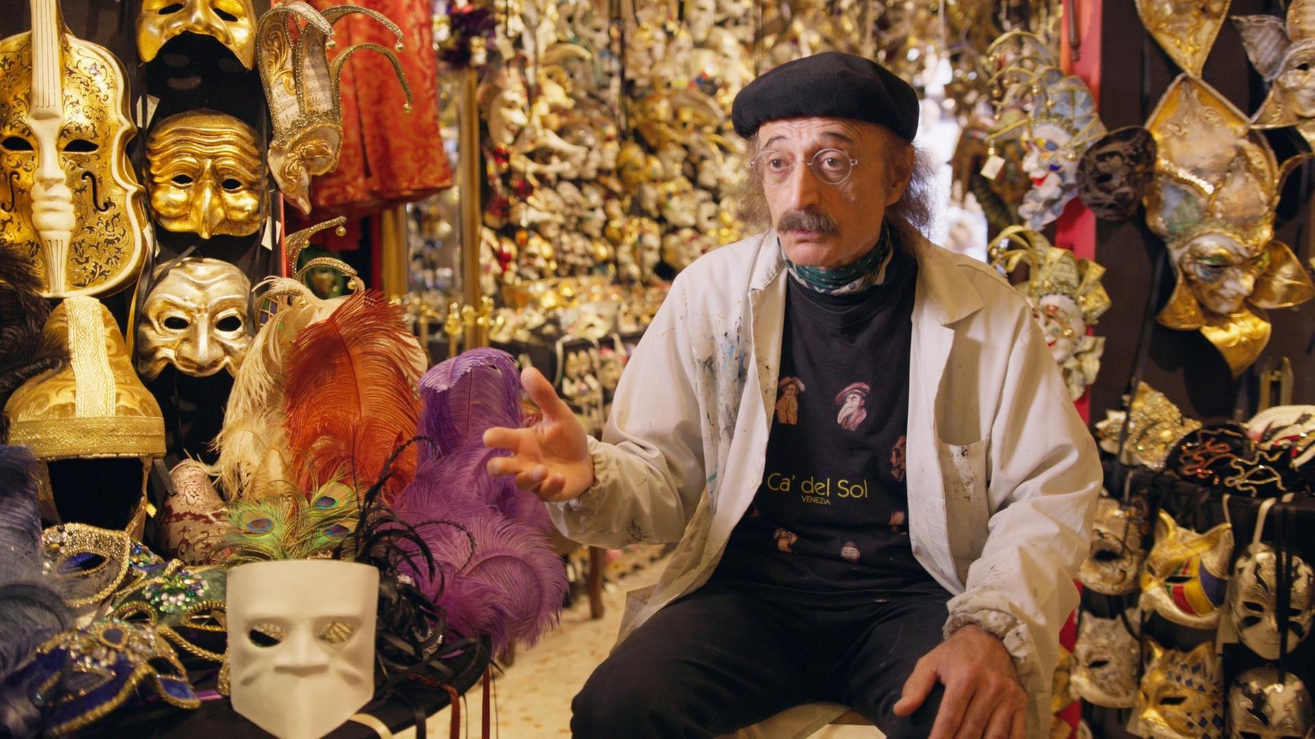 Ein Mann sitzt auf einem Stuhl in einem kleinen Raum voller venezianischer Masken.