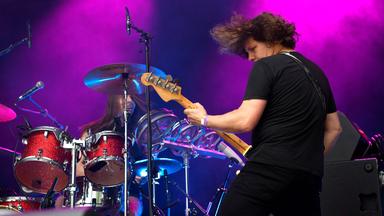 Auf einer lila angeleuchteten Bühne spielt ein Mann mit dem Rücken zum Publikum Gitarre, vor ihm sitzt ein Mann am Schlagzeug.