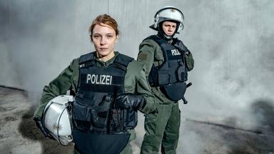 Eine Polizistin in Uniform. Im Hintergrund ein weiterer Polizist.