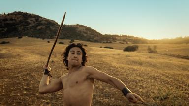 Ein junger Mann holt oberkörperfrei zu einem Speerwurf aus. Im Hintergrund ist eine steppenartige Landschaft zu sehen. 