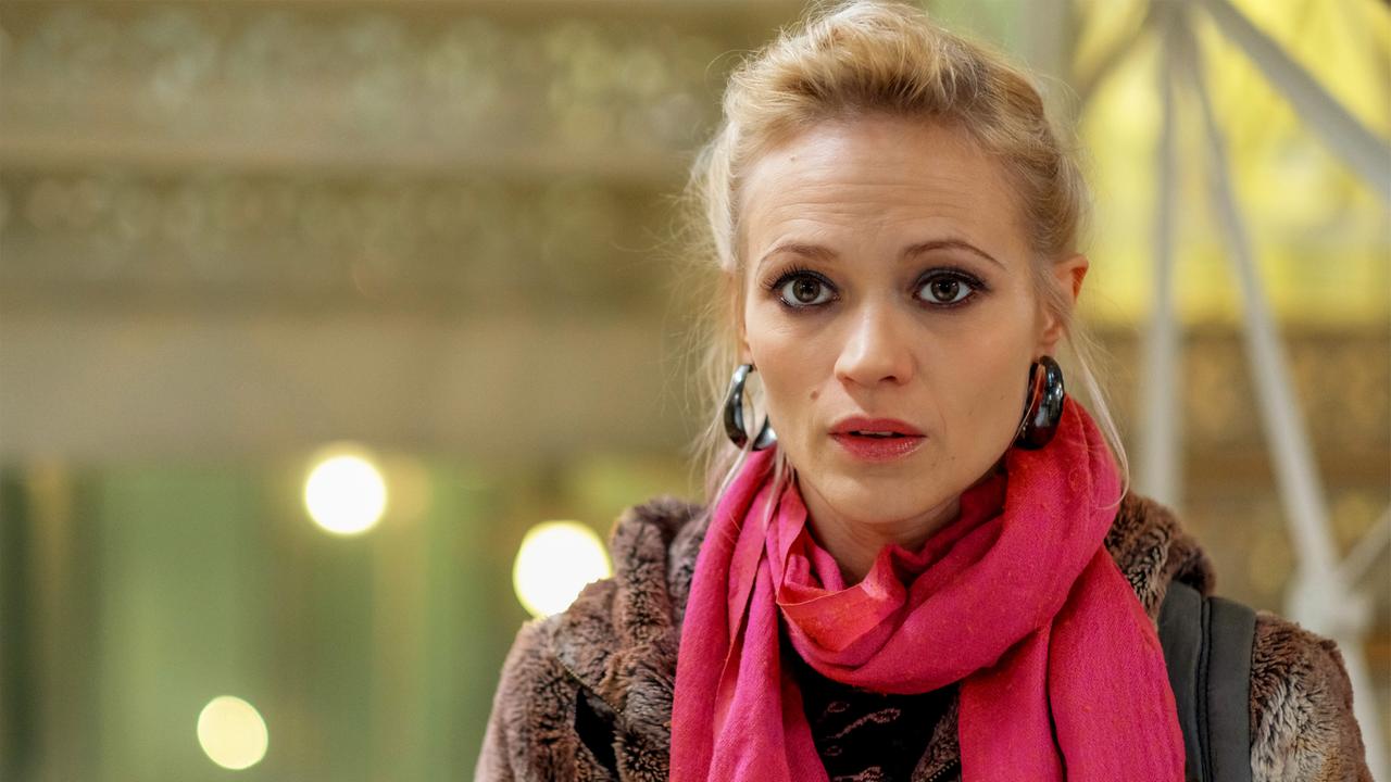 Im Bild ist eine junge Frau mit blonden Haaren und einem rosa Schal zu sehen. Sie trägt einen pelzartigen Mantel. Der Hintergrund ist unscharf und enthält weiche Lichtpunkte.