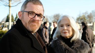 Das Bild zeigt zwei Personen in formeller Kleidung. Eine Frau mit grauen Haaren und einem dunklen Mantel trägt einen pelzigen Kragen. Ein Mann mit Brille hat einen Bart und schaut ernst. Im Hintergrund sind weitere Menschen und Bäume sichtbar.