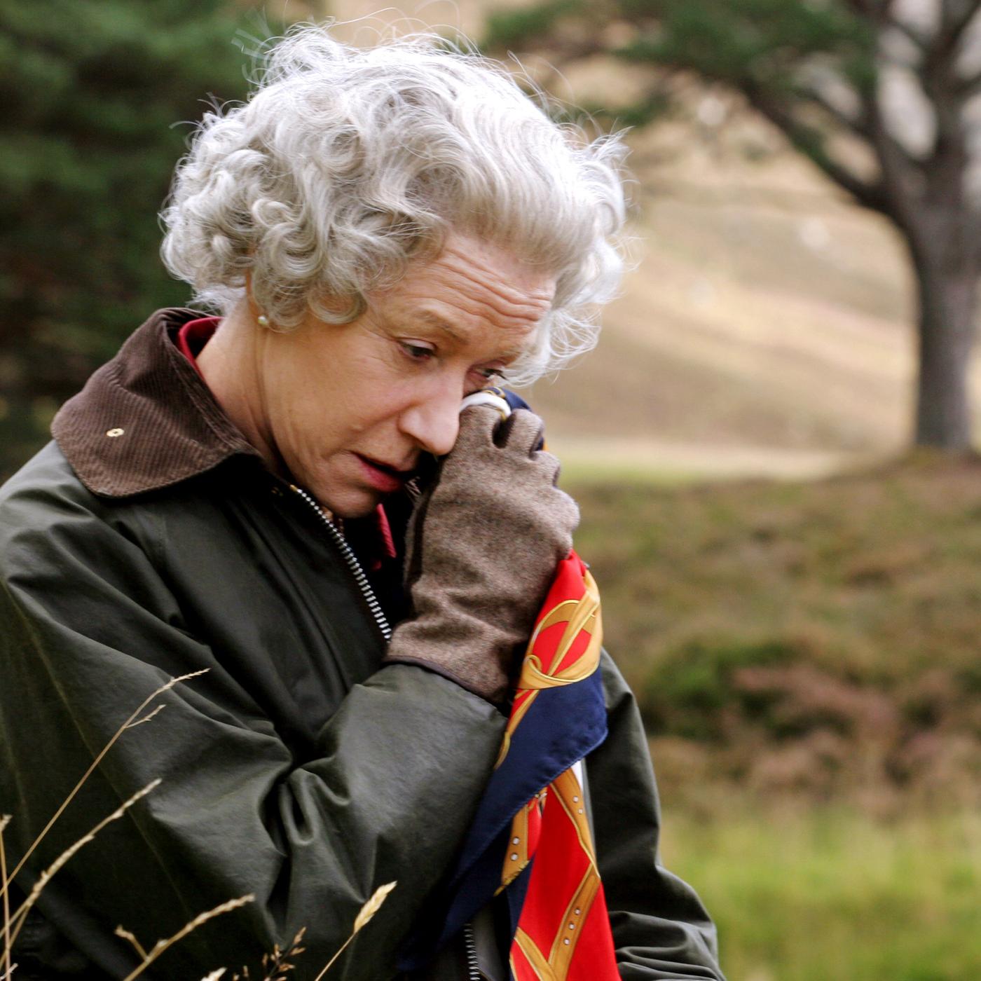 Vor dem unscharfen Hintergrund eines Baums in der Natur steht die Queen (Helen Mirren), die eine Wachsjacke mit Cordkragen und Lederhandschuhe trägt und mit gesenktem Kopf sich mit einem Seidentuch die Partie unter ihrem linken Auge betupft.