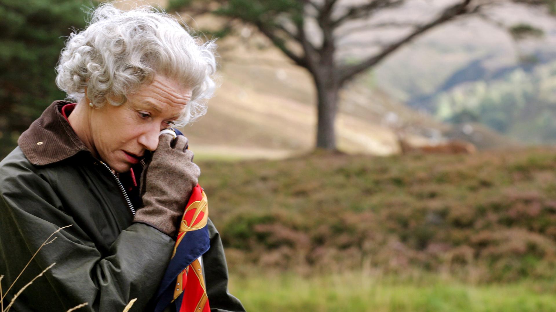 Vor dem unscharfen Hintergrund eines Baums in der Natur steht die Queen (Helen Mirren), die eine Wachsjacke mit Cordkragen und Lederhandschuhe trägt und mit gesenktem Kopf sich mit einem Seidentuch die Partie unter ihrem linken Auge betupft.