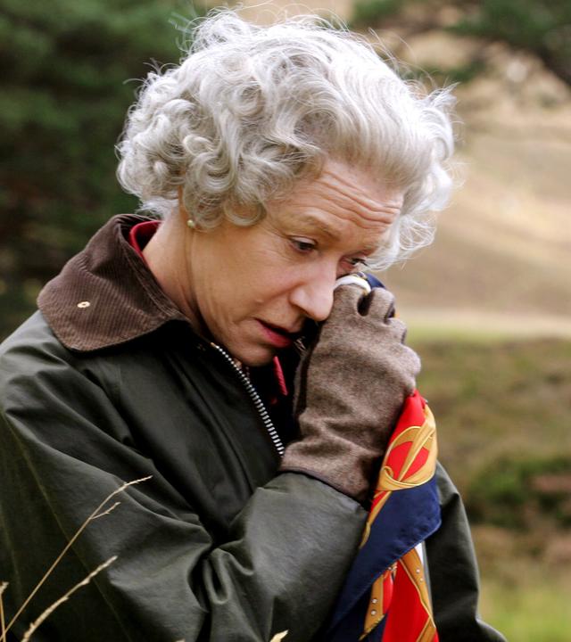 Vor dem unscharfen Hintergrund eines Baums in der Natur steht die Queen (Helen Mirren), die eine Wachsjacke mit Cordkragen und Lederhandschuhe trägt und mit gesenktem Kopf sich mit einem Seidentuch die Partie unter ihrem linken Auge betupft.