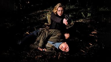 Eine Frau zielt mit einer Pistole rechts neben die Kamera. Neben ihr liegt ein verletzter Mann.