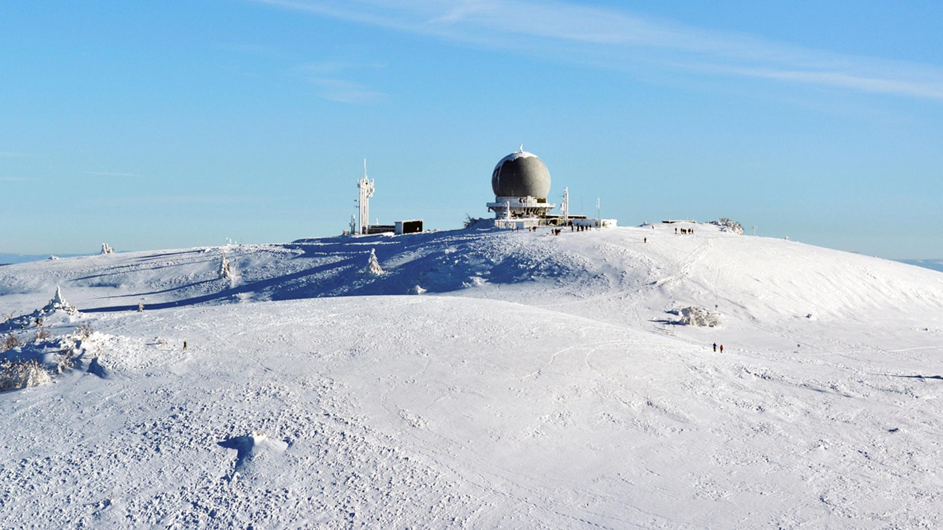 Ein mit Schnee bedeckter Hügel. Auf seiner Kuppe sind ein Gebäude und Antennen zu erkennen