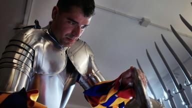 Ein Mitglied der Schweizer Garde trägt eine glänzende Rüstung und poliert ein Schild, während im Hintergrund Hellebarden zu sehen sind.