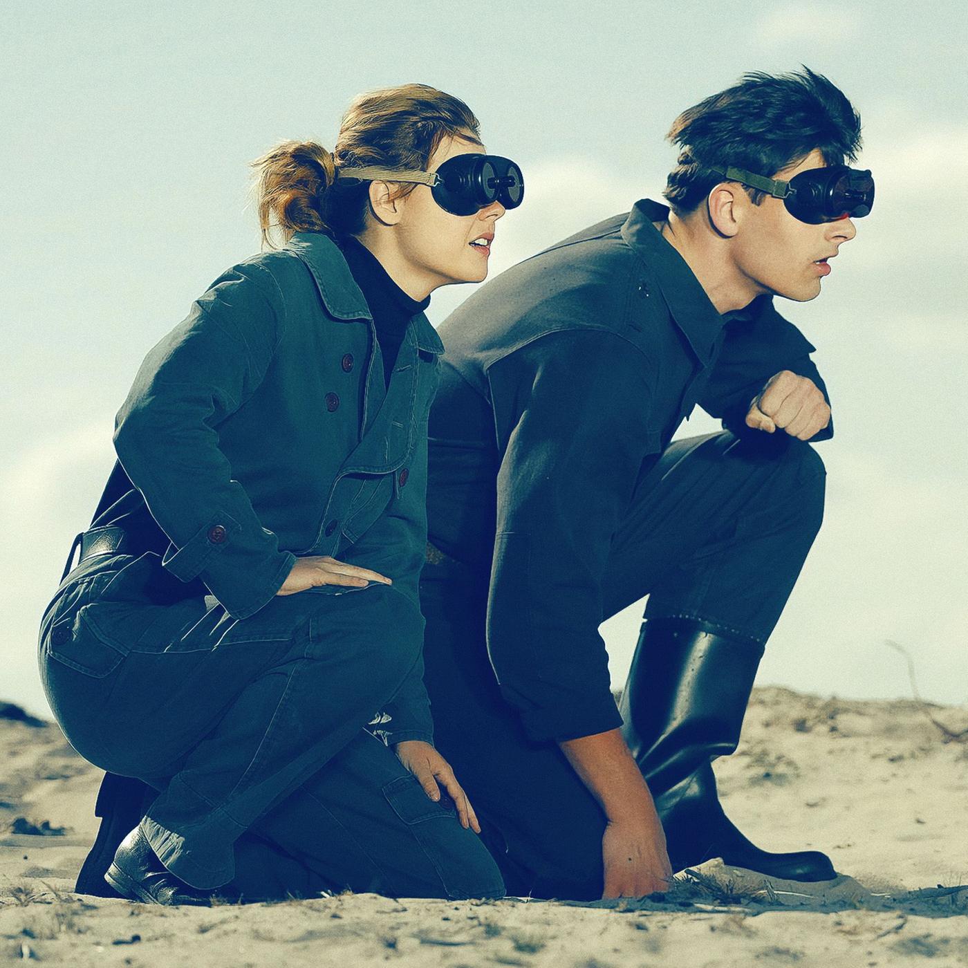 Eine junge Frau und ein junger Mann knien mit Brillen in einer Wüste.