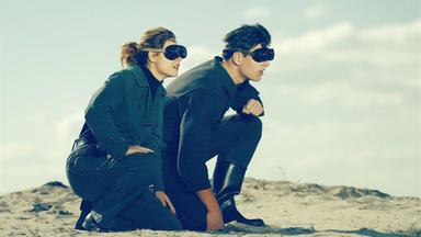 Eine junge Frau und ein junger Mann knien mit Brillen in einer Wüste.