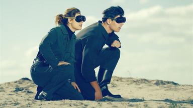 Eine junge Frau und ein junger Mann knien mit Brillen in einer Wüste.