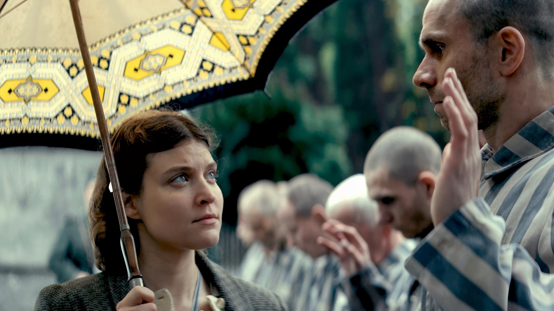 Eine Frau mit Schirm geht an Männern in gestreiften Häftlingsanzügen vorbei.