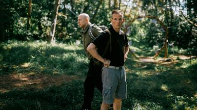 Zwei Personen befinden sich in einem bewaldeten Gebiet mit hohen Bäumen und üppigem Grün. Die Person im Vordergrund trägt ein schwarzes Shirt und graue Shorts, hat die Hände in die Hüften gestemmt und einen Rucksack auf dem Rücken. Die zweite Person steht etwas weiter hinten, trägt ein grünes Shirt und dunkle Hose und ebenfalls einen Rucksack. Sonnenlicht fällt durch die Bäume und erzeugt ein lebendiges Spiel aus Licht und Schatten auf dem Waldboden. Die Szene wirkt ruhig und natürlich und spielt sich offenbar tagsüber ab. 