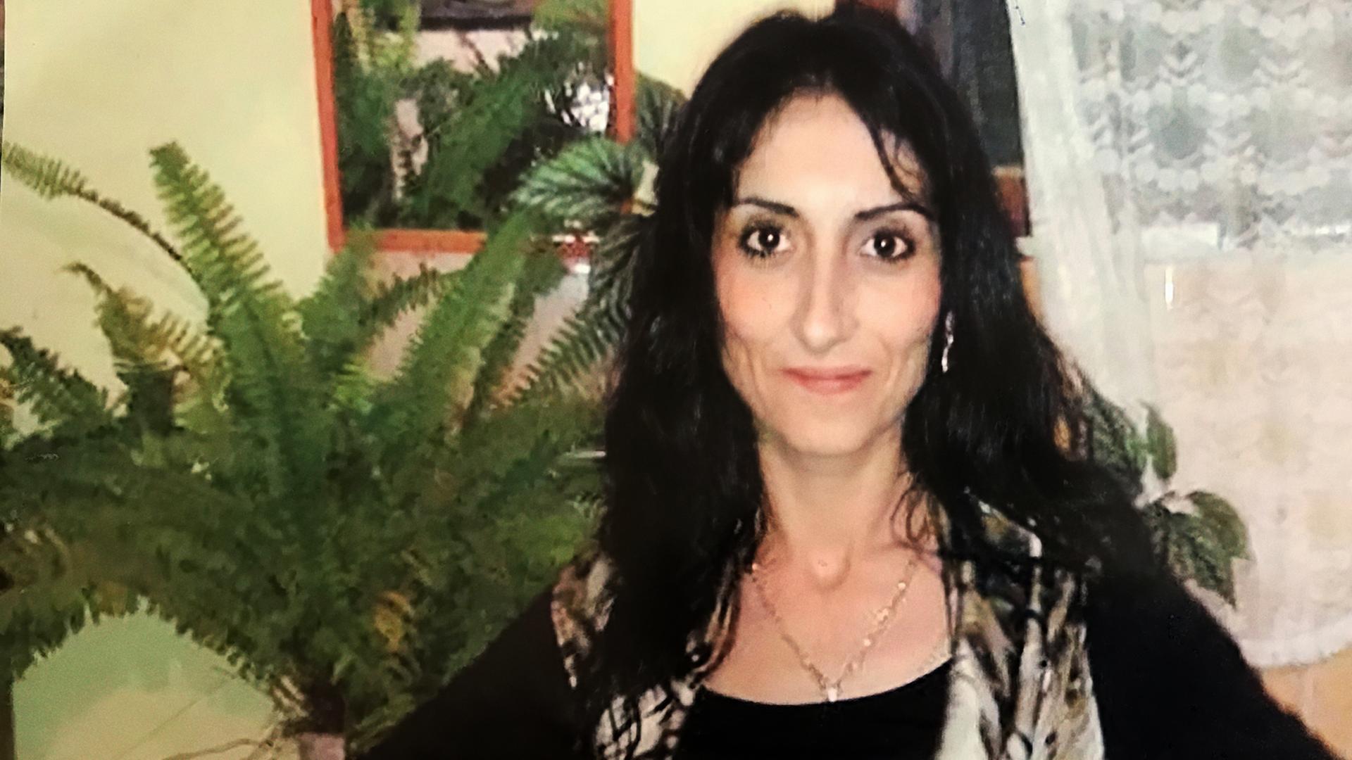 Eine Frau mit langen dunklen Haaren steht in einem Wohnzimmer vor einem Fenster mit Spitzengardinen. Im Hintergrund befinden sich mehrere große grüne Zimmerpflanzen und ein Spiegel.