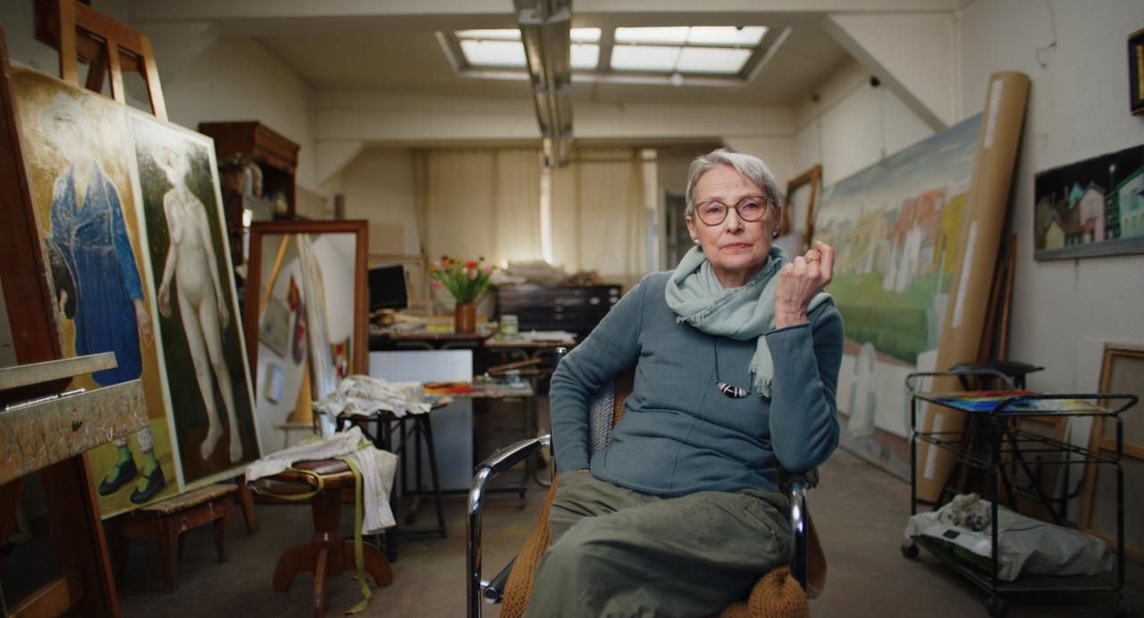 Eine Frau sitzt in der Mitte eines Atelierraums. Im Hintergrund sind verschiedene Gemälde zu sehen.