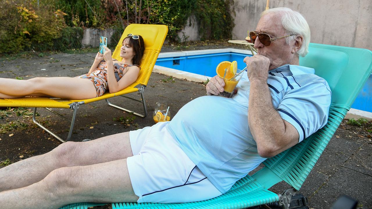 Ein alter Mann und eine junge Frau sitzen in einem Cabrio und fahren eine Landstraße entlang.