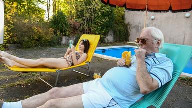 Eine junge Frau mit einem Buch und ein älterer Mann mit einem Cocktail liegen in einem Garten neben einem Pool auf Klappliegen und sonnen sich. 