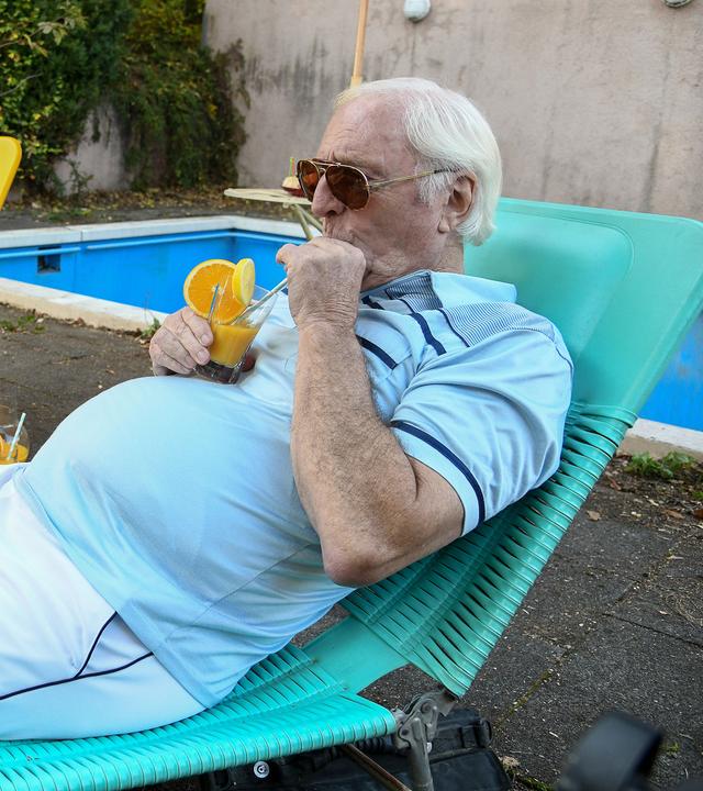 Eine junge Frau mit einem Buch und ein älterer Mann mit einem Cocktail liegen in einem Garten neben einem Pool auf Klappliegen und sonnen sich. 