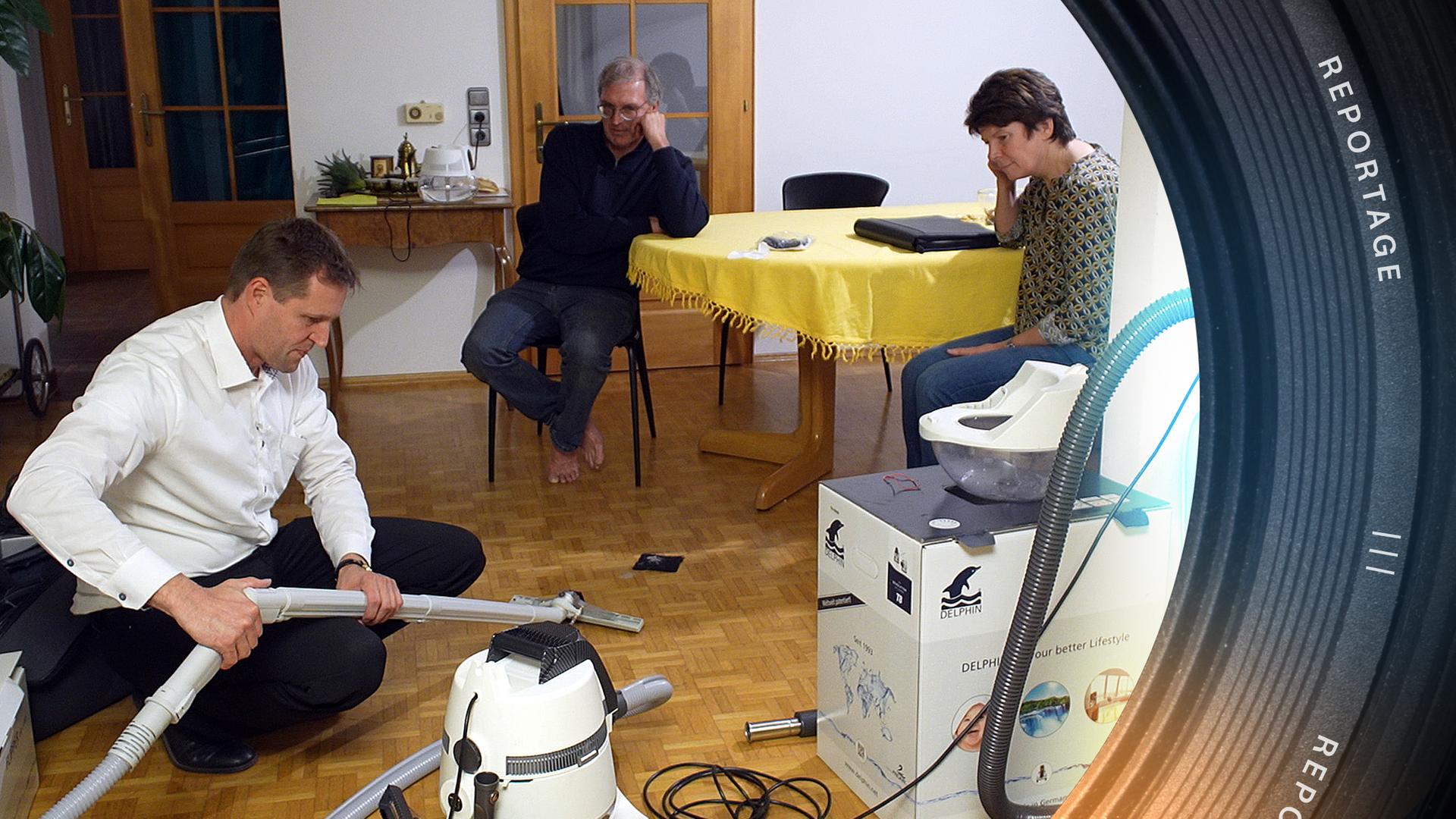 Bernhard Grassinger in der Hocke an einem seiner Staubsaugermodelle. Im Hintergrund am Tisch zwei Kunden, die ihm dabei zuschauen.