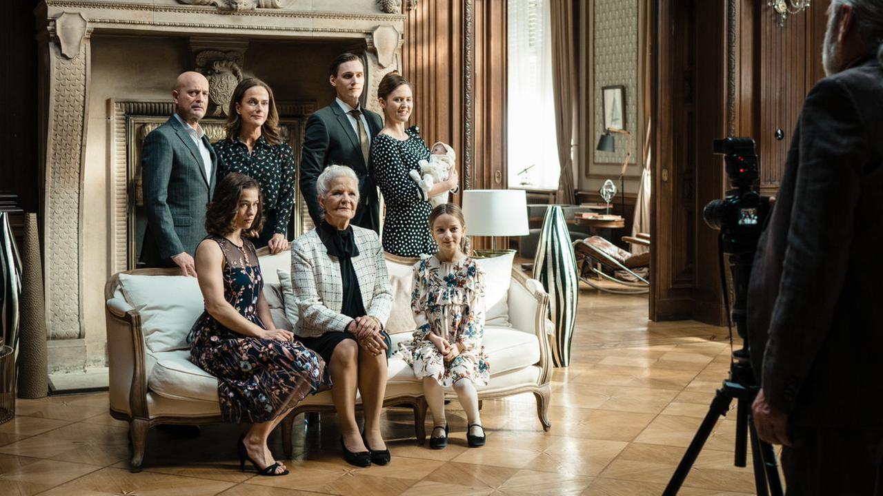 Die verlorene Tochter - Das Irrenhaus, das man Familie nennt": Familie von Gems hat sich für ein Familienfoto vor dem Kamin ihres Wohnzimmers versammelt: Heinrich (Christian Berkel), Isa (Henriette Confurius), Sigrid (Claudia Michelsen), Lore (Hildegard Schmahl), Philip (Rick Okon), Veronica (Emily Cox) und Celia (Una Lir). Ihnen gegenüber steht der Fotograf (Wolfgang Ikert), der von hinten zu sehen ist. Er schaut die Familie an, vor ihm steht eine Fotokamera auf einem Stativ.