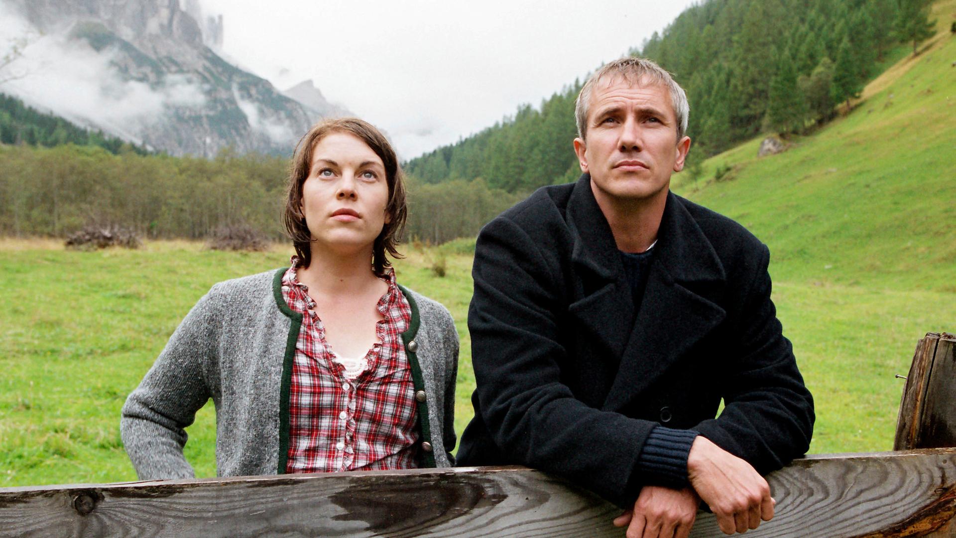 Benedikt (Markus Böker) und Johanna (Victoria Mayer) stehen in den Bergen auf einer Weide am Zaun und blicken nach vorne