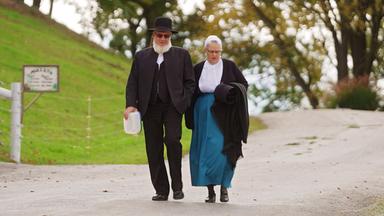 Ein älteres Paar in antiquierter Kleidung geht eine schmale Straße entlang.