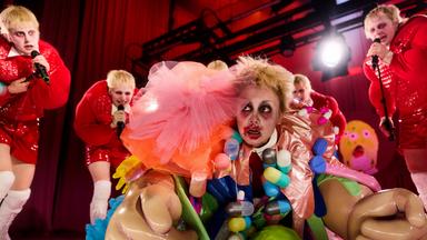 Unter dem herabgelassenen Bühnenvorhang krabbeln sechs gleich aussehende junge androgyne Personen mit kurzen blonden Haaren, rot-weißer Kleidung und stark geschminkten Augen hervor.