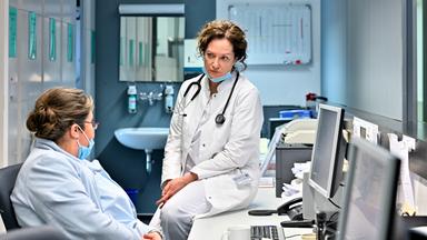 Eine Ärztin mit weißem Kittel und Stethoskop sitzt auf einem Schreibtisch und spricht mit einer Kollegin, die eine Maske trägt. Im Hintergrund sind Computerbildschirme und medizinische Unterlagen zu sehen.