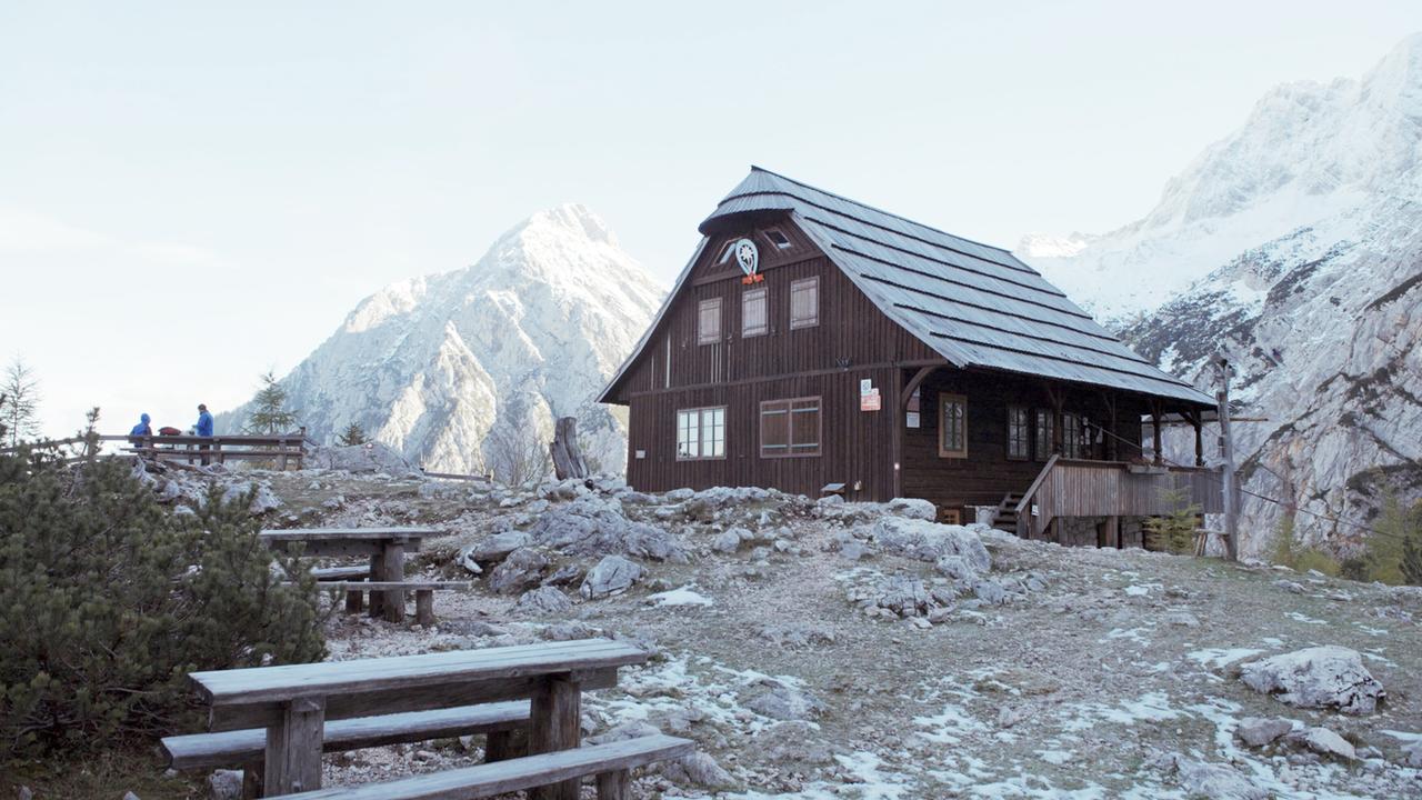 Eine Berghütte inmitten einer alpinen Landsschaft, im Vordergrund hölzerne Bänke und Tische