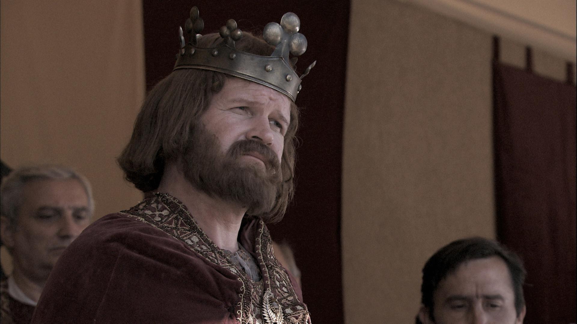 Ein König in historischer Gewandung und Krone, der von innerhalt eines Zeltes in die Ferne blickt.