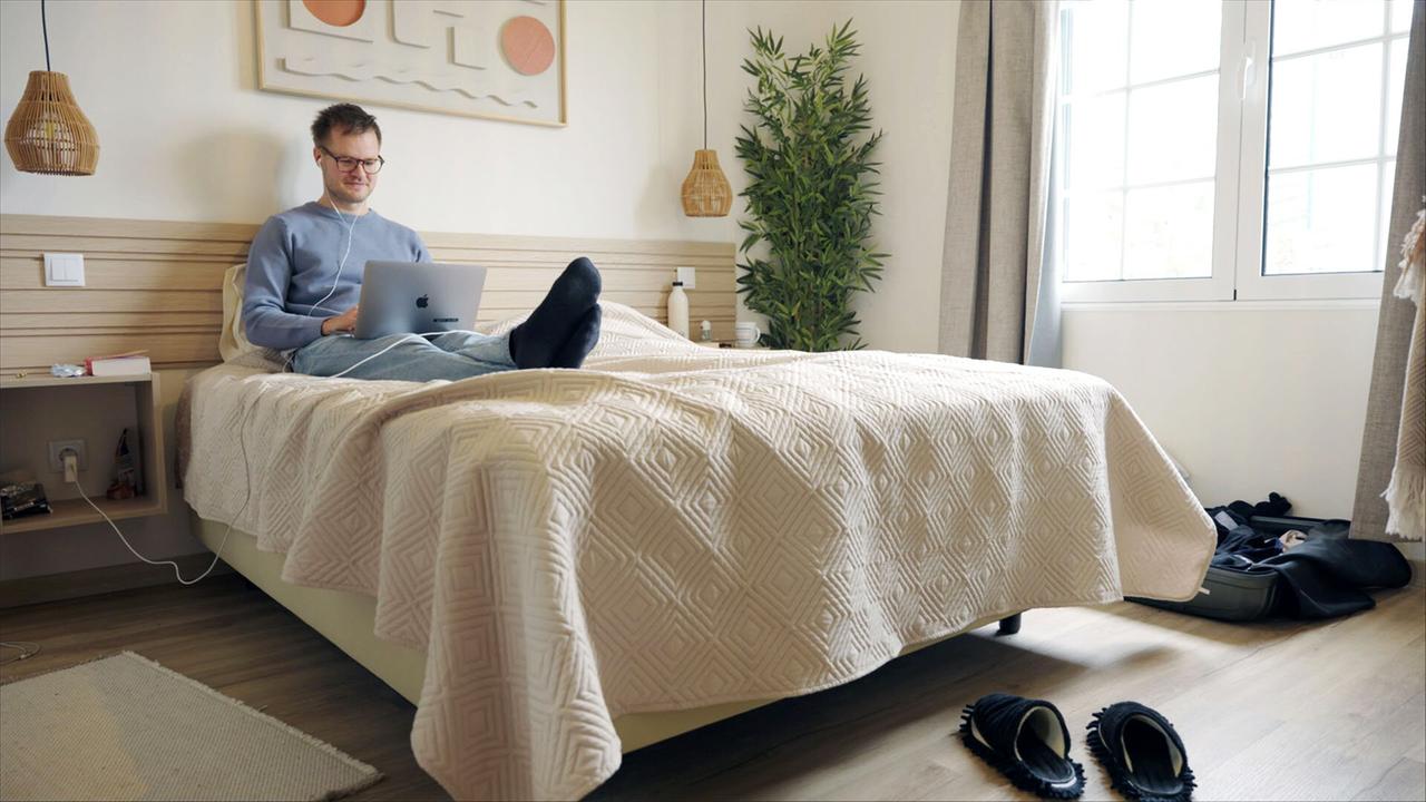 Das Bild zeigt einen Raum, der als Schlafzimmer dient. In der Mitte des Raumes steht ein Doppelbett, das mit einer hellen, strukturierten Tagesdecke bedeckt ist. Auf dem Bett sitzt ein Mann mit Brille und lächelndem Gesicht, der auf einem Laptop arbeitet. Er trägt einen hellblauen Pullover und hat die Beine auf dem Bett ausgestreckt.   Neben dem Bett steht ein kleines Nachttischchen auf der linken Seite, auf dem sich einige persönliche Gegenstände befinden. Neben dem Nachttisch führt ein Kabel zu einer Wandsteckdose, in der ein Gerät eingesteckt ist.   Im Hintergrund ist eine große Fensterfront zu sehen, die viel Tageslicht in den Raum lässt. Vor dem Fenster hängen zwei helle Gardinen. In der Ecke des Raumes steht ein grüner, kunstvoller Pflanze.   Der Boden ist aus hellem Holz und vor dem Bett liegt ein kleiner Teppich. Am Fußende des Bettes sind ein paar schwarze Hausschuhe und ein geöffnetes Gepäckstück zu sehen, das mit persönlichen Gegenständen gefüllt ist.   Die gesamte Atmosphäre des Raumes wirkt modern und freundlich, mit einem klaren Fokus auf Funktionalität und Komfort für eine digitale Lebensweise.