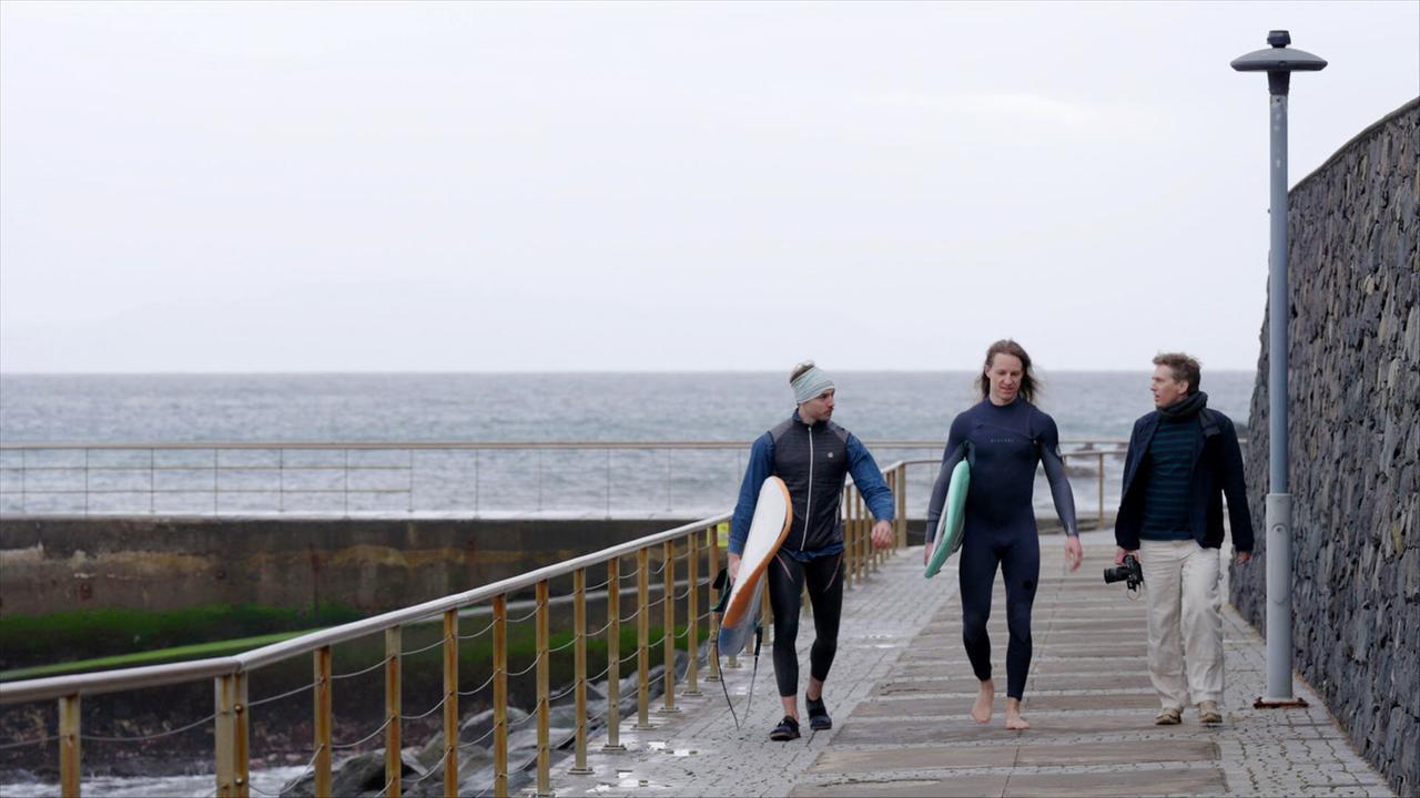Auf dem Bild sind drei Personen zu sehen, die auf einem Gehweg an der Küste von Madeira entlang gehen. Zwei der Personen tragen Wetsuits und halten Surfboards in der Hand. Eine Person hat ein braunes Surfboard, während die andere ein hellgrünes Surfboard trägt. Die dritte Person, die keine Taucherbekleidung trägt, hat einen schwarzen Schal um den Hals und trägt eine Kamera.   Im Hintergrund erstreckt sich das Wasser des Atlantischen Ozeans, das eine graue Farbe hat, und der Himmel ist bewölkt. Auf der rechten Seite des Gehwegs steht eine Beleuchtungssäule. Der Gehweg selbst besteht aus grauen Steinen und betont einen maritimen, aktiven Lebensstil. Die Szenerie vermittelt eine entspannte Atmosphäre, die mit dem Surfen assoziiert wird.