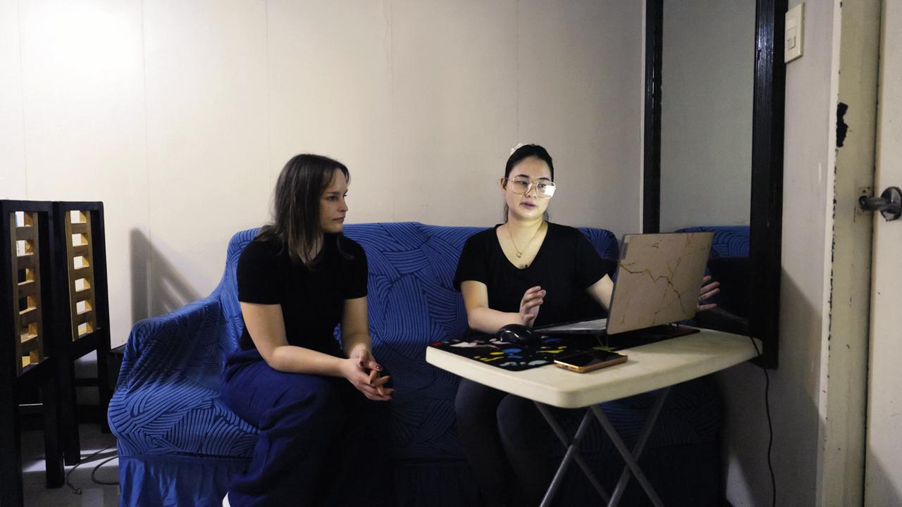 Auf dem Bild sind zwei Frauen in einem Raum zu sehen, der schlicht möbliert ist. Eine Frau sitzt auf einem blauen Sofa mit einer gestreiften Decke, während die andere Frau neben ihr auf einem Stuhl sitzt. Beide tragen schwarze Oberteile. Die Frau am Tisch zeigt auf einen Laptop, der auf einem kleinen weißen Tisch steht. Auf dem Tisch liegt auch eine Maus und ein Smartphone. Der Raum hat helle Wände und einen großen Spiegel, der das Licht reflektiert. Der gesamte Raum wirkt etwas nüchtern und hat eine intime Atmosphäre.